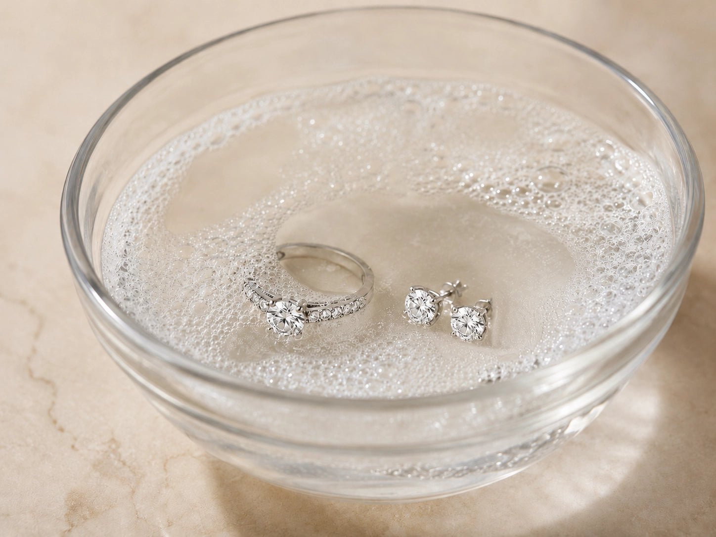 Close-up of diamond jewelry soaking in mild soapy water, illustrating a gentle at-home cleaning method for rings and earrings.