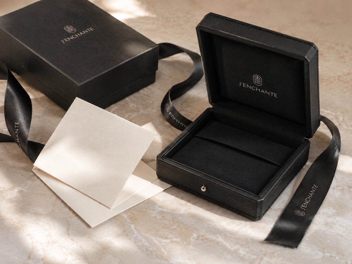 J'ENCHANTE gift box, jewelry case, ribbon, and card on a light surface, representing luxury jewelry gifting and packaging.